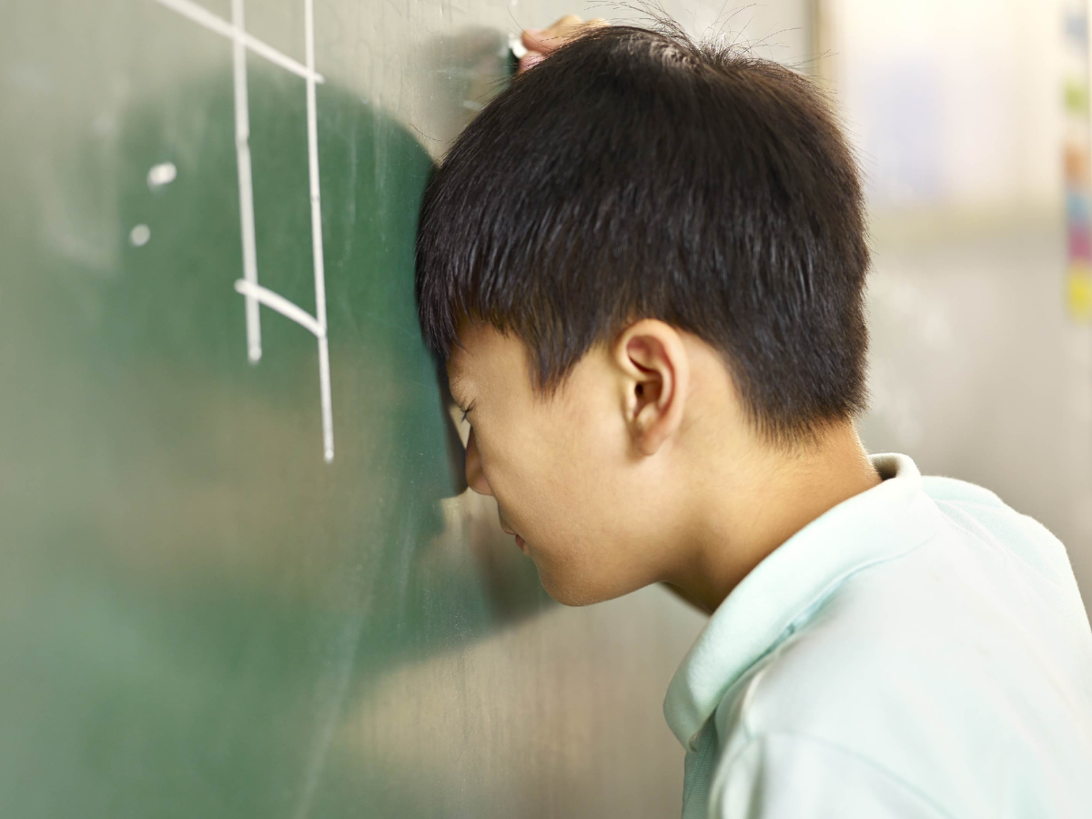 Seorang siswa laki-laki tampak menundukkan kepala ke papan tulis hijau dengan ekspresi frustrasi, mencerminkan tekanan akademis yang dialami di sekolah.