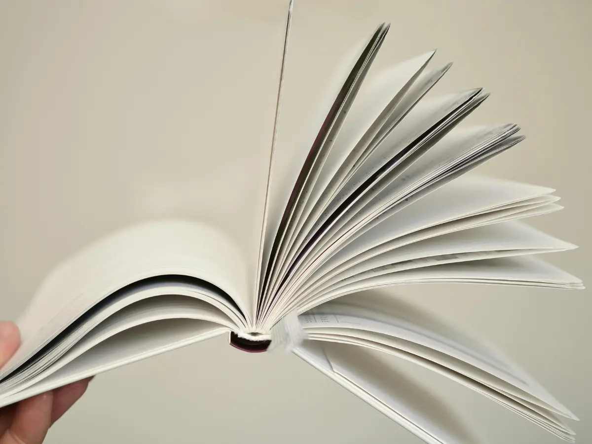 menampilkan buku yang terbuka lebar digenggam oleh tangan sebelah kiri dengan lembaran-lembarannya yang naik keatas difoto dari jarak dekat