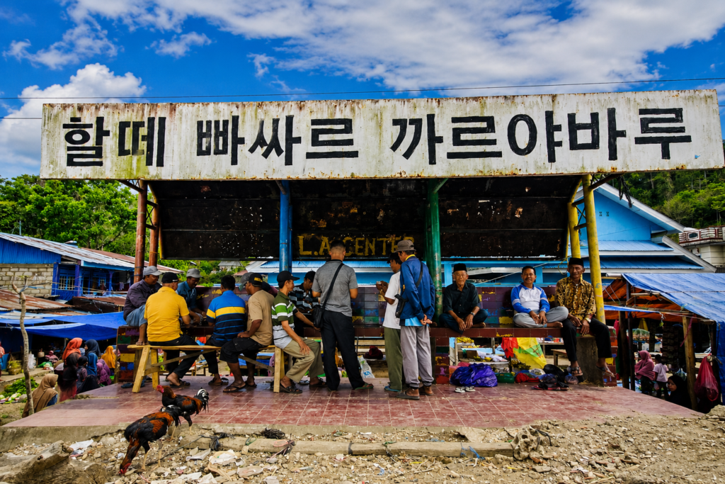 Halte di kawasan Pasar Karya Baru Baubau dengan tulisan Hangeul besar di atasnya dan warga beraktivitas di bawahnya.